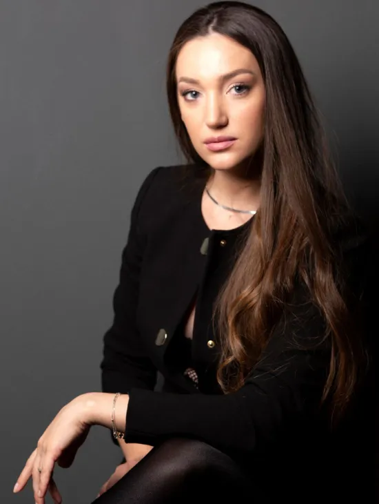 Enkeleda Kamani sitzt vor einem grauen Hintergrund und blickt in die Kamera. Sie trägt eine schwarze Jacke und hat lange braune Haare, die über eine Schulter fallen.