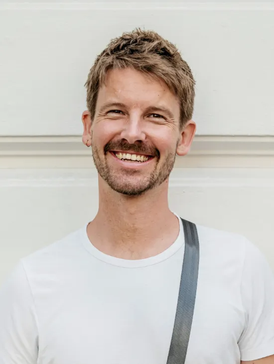 Josua Guss mit kurzen braunen Haaren und Bart lächelt in die Kamera. Er trägt ein weißes T-Shirt und einen grauen Schultergurt vor einer hellen Wand.