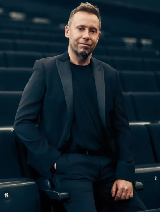 Dirigent Matteo Beltrami in schwarzem Anzug und schwarzem Shirt zwischen den Sitzreihen eines Zuschauerraums. Er hat eine Hand in der Hosentasche und blickt in die Kamera.