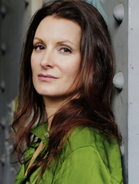 Porträt von Nadine Schori mit langem, dunkelbraunem Haar und grüner Bluse. Sie lehnt an einer grauen Metallkonstruktion mit großen Nieten und blickt direkt in die Kamera.