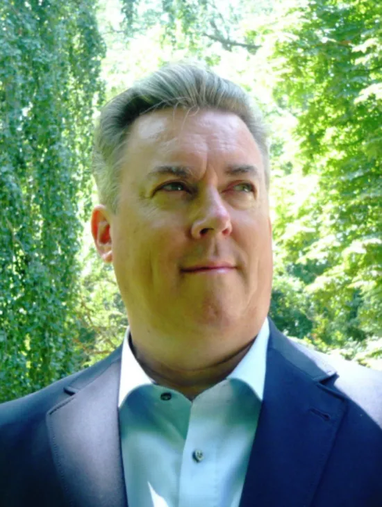Yves Saelens trägt ein dunkelblaues Sakko und ein hellblaues Hemd. Er blickt leicht lächelnd zur Seite; im Hintergrund sind grüne Bäume zu sehen.