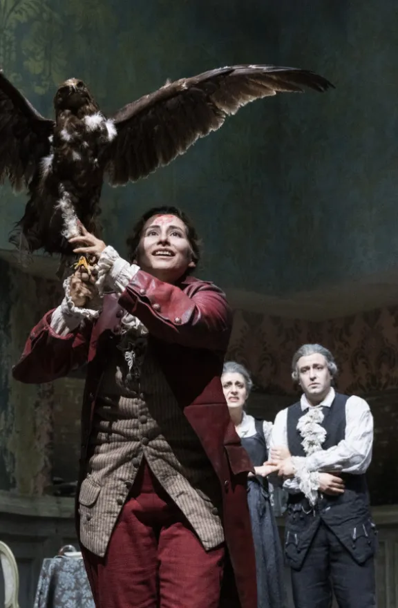 Cain (Adriana Bastidas-Gamboa), dressed in a red Baroque costume, holds a large eagle with outstretched wings on his arm and looks upwards with a smile. In the background, Eve (Tamara Banješević) and Adam (Dmitry Ivanchey) watch the scene. Scene from Alessandro Scarlatti’s oratorio “Cain and Abel”.