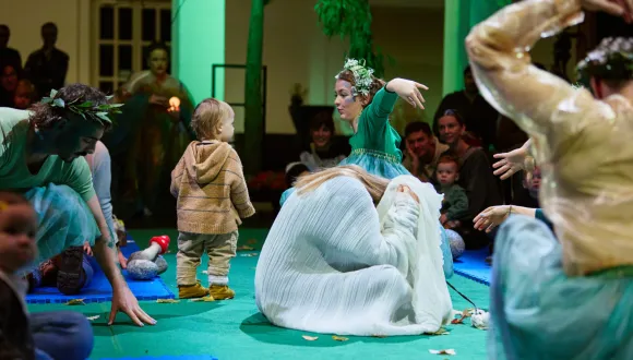 Scene from the opera for very young children Everything That Flies. In a circular performance space, a toddler stands among several performers wearing flowing costumes in shades of green, blue, and white. One performer, wearing a wreath of leaves, gestures expressively toward the child. Parents and children sit around the performance area, experiencing the action at close range. Natural elements, fabrics, and scattered leaves create a poetic and playful atmosphere.