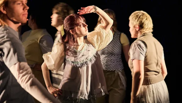 Scene from the performance When I Find Myself in Times of Trouble by the Opera, Song and Scene theatre club: Several young adults move across a dimly lit stage. At the center stands a person with auburn hair, shielding their eyes with one hand as if looking into the distance or searching for direction. Warm stage lighting highlights the central figure, while other participants pass by in different directions. The scene conveys movement, searching, and self-reflection.