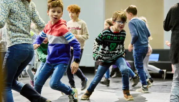 A school class is actively moving through a large indoor space. Several children are running, jumping, and interacting with one another in a playful group exercise. In the foreground, two children can be seen in dynamic motion, while other pupils participate in the background. The scene conveys energy, teamwork, and collaborative learning through movement.