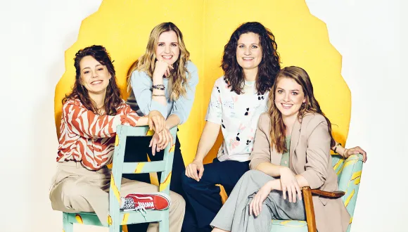 Group portrait of the Oper.Stadt.Schule music theatre education team at Oper Köln. Four women—Nadine Syskowski, Katja Fischer, Stephanie Sonnenschein (Head of Department), and Anna Mangold—are arranged in a bright studio setting against a decorative, glowing yellow backdrop. Two are seated on brightly designed chairs, while the other two stand or lean against them. All are smiling at the camera. Their relaxed poses and the colourful elements create an open, creative, and welcoming impression.