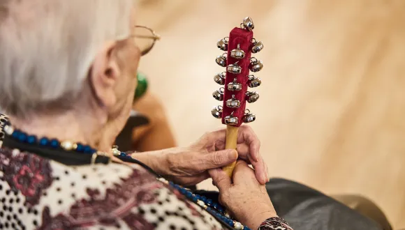 An older woman attends an Oper.Stadt.Schule event and holds a handheld instrument with numerous small bells. The photograph is taken from behind, focusing on her hands and the instrument. Her calm, attentive posture conveys active music-making, participation, and the joy of experiencing music together.