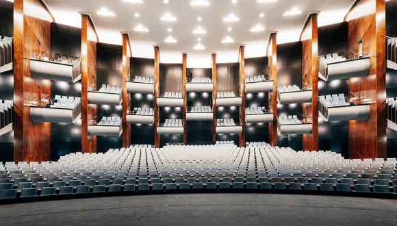Wide-angle view of the large auditorium of the Cologne Opera at Offenbachplatz, featuring several tiers stacked above one another. The light-colored rows of seats fill the space evenly, while vertical wooden panels and dark wall surfaces structure the architecture.