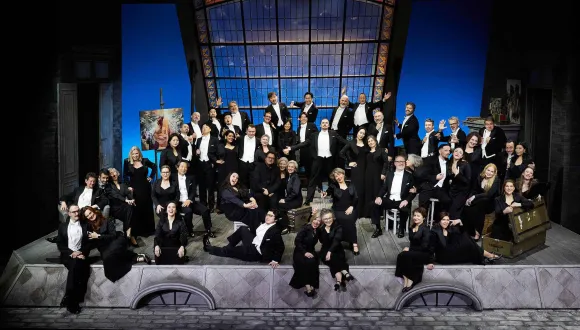 Group portrait of the Cologne Opera Chorus on the opera stage. More than 50 singers are posing in elegant black concert attire in a variety of seated and standing positions. In the background, a large window revealing a starry night sky dominates the set, alongside stage elements including doors, a painting, and various props. Their relaxed poses convey a strong sense of ensemble and stage presence.