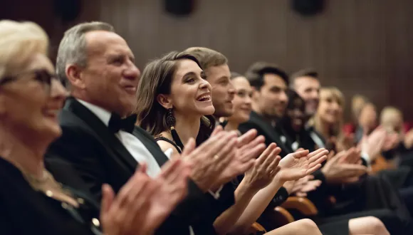 Several formally dressed audience members are seated in the auditorium of the Cologne Opera, applauding. At the centre, a woman in black evening wear smiles while the people around her enthusiastically applaud. The image conveys the festive atmosphere of an evening at the opera.