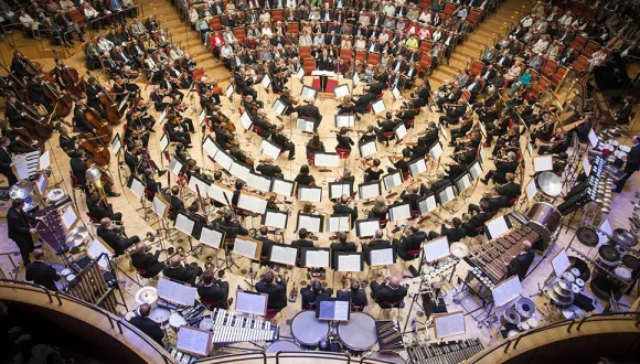 Top-down view of the Cologne Philharmonie concert hall: the Gürzenich Orchestra Cologne is seated in a large circular formation around the conductor’s podium. An extensive percussion section is prominently visible in the foreground. The stalls and surrounding tiers are filled with audience members. The image conveys the scale of the orchestra and the distinctive architecture of the concert hall.