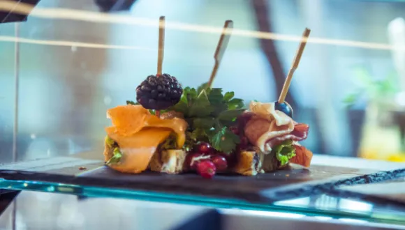 Close-up of a display featuring small open sandwiches topped with smoked salmon, ham, herbs, berries and cocktail picks. The food is presented on dark serving platters inside a glass display case, conveying the high quality of the culinary offering.