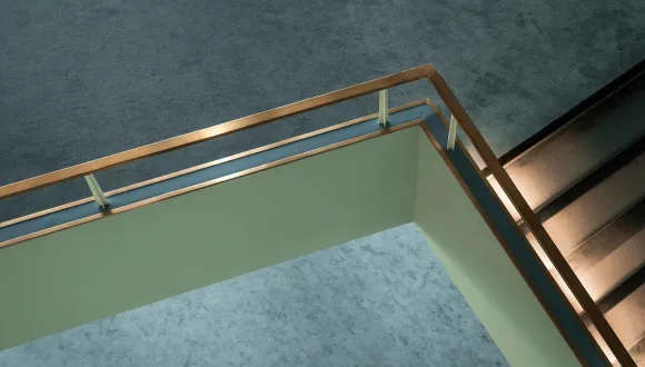 View of the stairwell at the Cologne Opera at Offenbachplatz. A light-coloured gallery with a brass handrail forms a striking angle beneath a dark textured ceiling. An illuminated staircase runs alongside it, highlighting the building's clean, modern architecture.