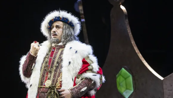 Der alte König Detlef (Ferhat Baday) hebt seine mit Ringen geschmückte Faust. Er trägt ein rot-goldenes Gewand mit weißem Fellbesatz, eine Krone und eine graue Perücke mit Bart. Szene aus der Kinderoper „DER KÖNIG VERSCHENKT WAS“ von Philipp Matthias Kaufmann.