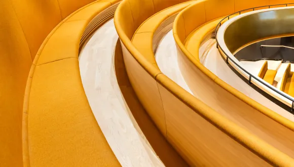 View of the balcony at Cologne Children’s Opera, with curved rows of yellow upholstered seating, light wooden floors and a parapet with a black railing.