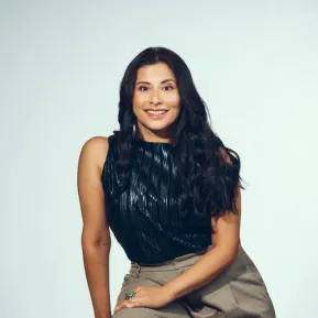 Mezzo-soprano Adriana Bastidas-Gamboa is seated on a black stool against a light grey studio background. She looks directly at the camera with a warm smile. Her long, dark, wavy hair falls over her shoulders. She wears a dark, subtly shimmering sleeveless top and wide-legged grey trousers. A large ring and hoop earrings stand out as distinctive accessories. The portrait conveys an open, confident, and approachable impression.