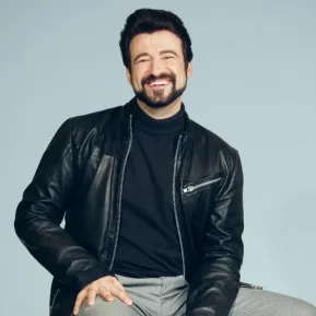 Tenor Dmitry Ivanchey is seated on a stool, smiling warmly at the camera. He is wearing a dark turtleneck sweater, a black leather jacket, and light grey trousers. His dark hair and neatly groomed beard give the portrait a distinctive appearance. Set against a light blue background, the photograph has a friendly, modern, and professional look.