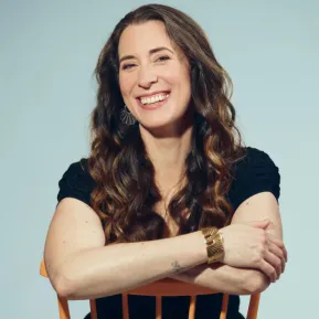 Soprano Emily Hindrichs sits behind a light wooden chair, resting her arms on the backrest. She smiles warmly at the camera. Her long, wavy brown hair falls over her shoulders. She is wearing a dark dress, distinctive earrings, and a wide bracelet. Set against a light blue background, the portrait has a friendly, relaxed, and professional appearance.