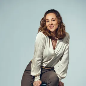 Soprano Giulia Montanari sits relaxed on a black stool against a light grey studio background. She leans slightly forward, smiles at the camera, and wears a cream-coloured, subtly lustrous long-sleeved blouse paired with grey trousers. Her shoulder-length, softly wavy hair frames her face naturally. The portrait conveys an open and friendly impression.