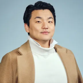 Baritone Insik Choi looks directly into the camera. He is wearing a light-coloured turtleneck sweater beneath a beige coat. His dark hair is combed back. Set against a light blue background, the portrait has a calm and professional appearance. The photograph shows him from the upper body upwards.