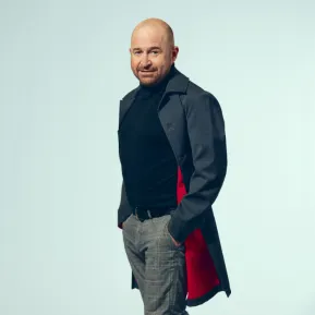 Tenor John Heuzenroeder stands slightly turned towards the camera, looking at the viewer with a subtle smile. He is wearing a dark turtleneck sweater, grey checked trousers, and a dark coat with a red lining. His hands are tucked into his trouser pockets. Set against a light blue background, the portrait has a modern and professional appearance. The photograph shows him in an almost full-length pose.