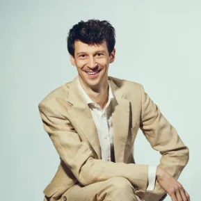 Baritone Wolfgang Stefan Schwaiger is seated on a light-coloured stool against a light grey studio background. He looks directly into the camera with a smile and wears a light beige suit with a white shirt. His legs are crossed, and his arms rest casually in front of him. His dark, slightly wavy hair and distinctive moustache are defining features of his appearance. The portrait conveys an open, friendly, and confident impression.