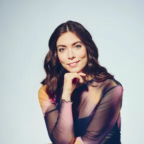 Portrait of soprano Alina König Rannenberg against a light grey studio background. She is seated at a slight angle to the camera, resting her chin on her right hand and looking directly into the lens with a warm smile. She is wearing a semi-transparent, long-sleeved dress in muted shades of violet, brown, and rose. Her shoulder-length dark brown hair falls in soft waves. Her relaxed pose conveys elegance and confidence.