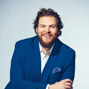 Portrait of bass Christoph Seidl against a light grey studio background. He is seated, leaning slightly forward, smiling directly at the camera, and wearing a bright blue suit with a white shirt and a patterned pocket square. His curly brown hair and full beard are defining features of his appearance. His relaxed posture conveys openness and confidence.