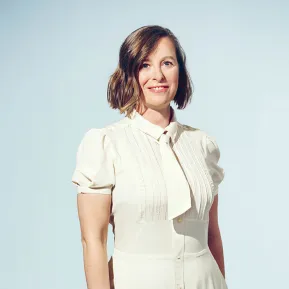 Portrait of soprano Claudia Rohrbach against a light grey studio background. She is standing at a slight angle, looking directly into the camera with a warm smile. She is wearing a cream-white dress with short puff sleeves, fine pintucks, and a bow at the collar. Her chin-length, slightly wavy brown hair is parted to one side. Her upright posture conveys elegance and composure.