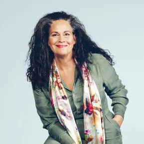 Portrait of Dalia Schaechter. The mezzo-soprano smiles at the camera while sitting slightly leaning forward. She wears an olive-green trouser suit with a long, colourful patterned scarf. Her long, curly dark hair falls loosely over her shoulders. The background is light blue and uncluttered.