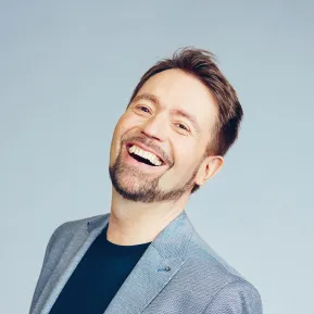 Portrait of Kammersänger Miljenko Turk. The baritone smiles with his mouth open and looks slightly upward. He wears a light blue blazer over a dark T-shirt. His short brown hair is styled back, and he has a neatly trimmed beard. The background is light blue and uncluttered.