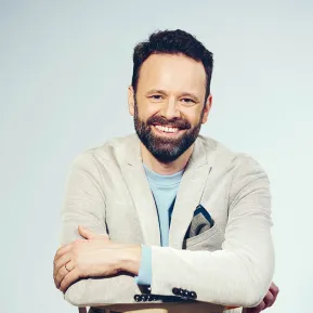 Portrait of Lucas Singer. The bass smiles directly at the camera while resting his folded arms on the back of a light wooden chair. He wears a light grey blazer over a light blue shirt. His dark hair is cut short, and he has a neatly trimmed full beard. The background is light blue and uncluttered.