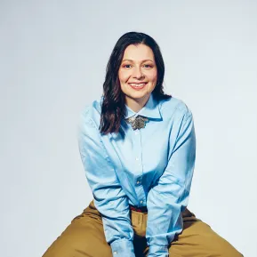 Portrait of soprano Maike Raschke against a light grey studio background. She is seated on a stool, leaning slightly forward and smiling directly at the camera. She is wearing a light blue blouse with a distinctive brooch at the collar and wide beige trousers. Her shoulder-length dark hair falls loosely over her shoulders. Her open posture conveys friendliness and natural ease.