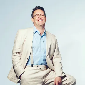 Portrait of Martin Koch. The tenor smiles with his mouth open and looks slightly upward. He wears a light-coloured linen suit with a light blue shirt, sits casually on a black stool, and has one hand in his trouser pocket. His bright pink socks stand out. The background is light blue and uncluttered.