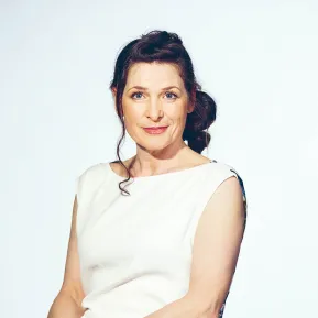 Portrait of Regina Richter. The mezzo-soprano sits with her hands loosely folded and looks into the camera with a gentle smile. She wears a sleeveless white dress with a subtly patterned back panel. Her dark hair is styled in a loose bun. The background is light blue and uncluttered.