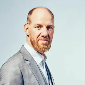 Portrait of Tijl Faveyts. The bass looks directly at the camera with a subtle smile. He wears a light grey blazer over a white shirt and a grey waistcoat. His reddish-blond beard is neatly trimmed, and his hair is cut very short. The background is light blue and uncluttered.