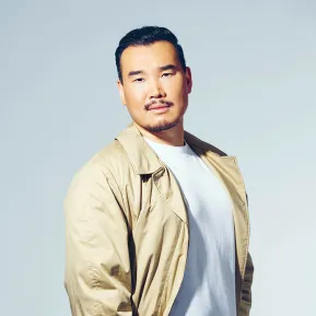 Portrait of tenor Young Woo Kim against a light grey studio background. He is standing at a slight angle to the camera, looking directly into the lens with a calm expression. He is wearing a beige coat over a white T-shirt and dark trousers. His short black hair, moustache, and goatee are defining features of his appearance. His relaxed posture, with one hand in his trouser pocket, conveys composure and confidence.