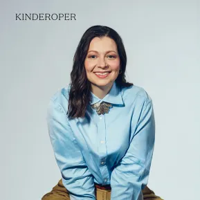Portrait of soprano Maike Raschke against a light grey studio background. She is seated on a stool, leaning slightly forward and smiling directly at the camera. She is wearing a light blue blouse with a distinctive brooch at the collar and wide beige trousers. Her shoulder-length dark hair falls loosely over her shoulders. Her open posture conveys friendliness and natural ease.